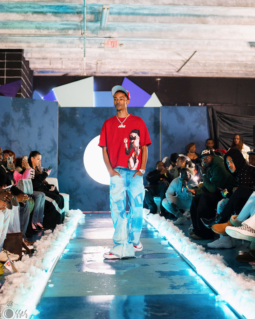 Model on a runway wearing a red shirt and light blue pants, with an audience and colorful background.